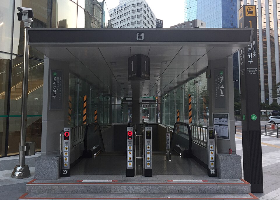 A street-level subway station entrance with escalators leading underground, flanked by modern glass office buildings in Seoul.