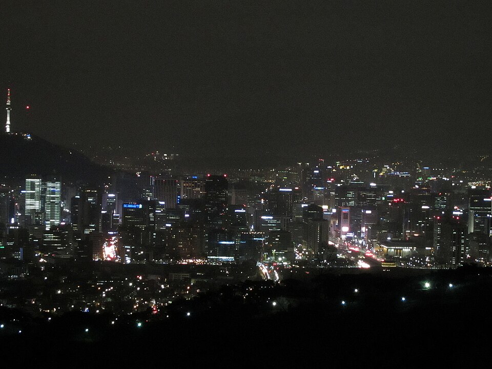A nighttime aerial view of Seoul's skyline showing illuminated skyscrapers with Namsan Tower visible on a hill in the background.