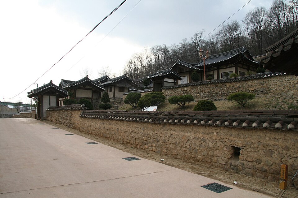 A row of traditional Korean tile-roofed houses sits behind a low stone wall on a wooded hillside beside a paved road.