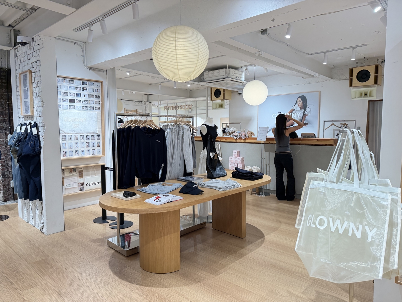 Interior of a clothing boutique with racks of dark garments, tote bags printed with the store's name, and a customer at the checkout counter.