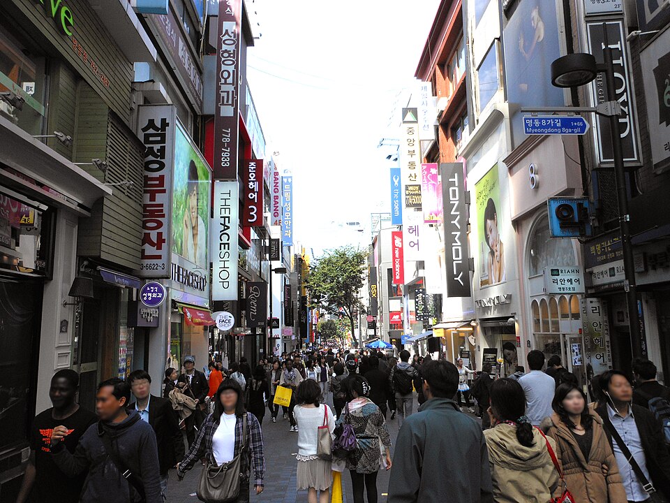 A crowded daytime street in Seoul's Myeong-dong shopping district lined with clothing and cosmetics stores and covered in colorful signage.