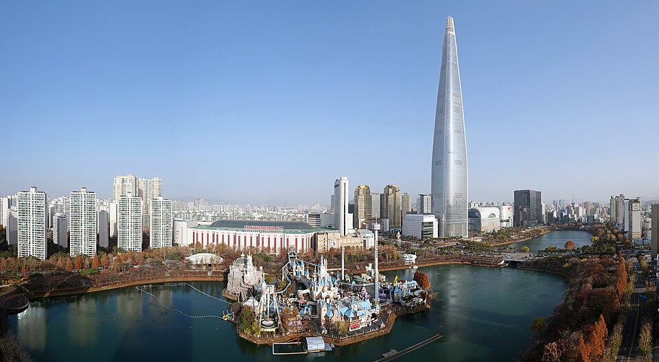 A daytime panoramic view of the tall, tapering Lotte World Tower rising above the Lotte World amusement park and Seokchon Lake in Jamsil, Seoul.