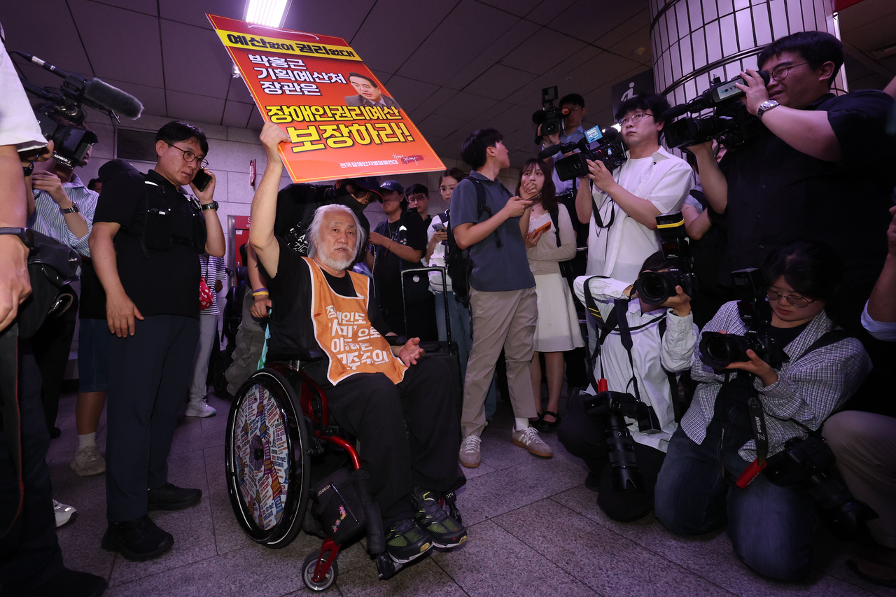 An elderly man in a wheelchair, wearing an orange vest, holds up a red placard with Korean text while surrounded by photographers and journalists in a subway station corridor.