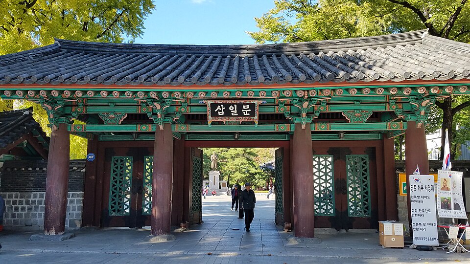 A traditional Korean wooden gate with a green tiled roof and Korean inscription stands at the entrance to Tapgol Park, with a person walking through it.