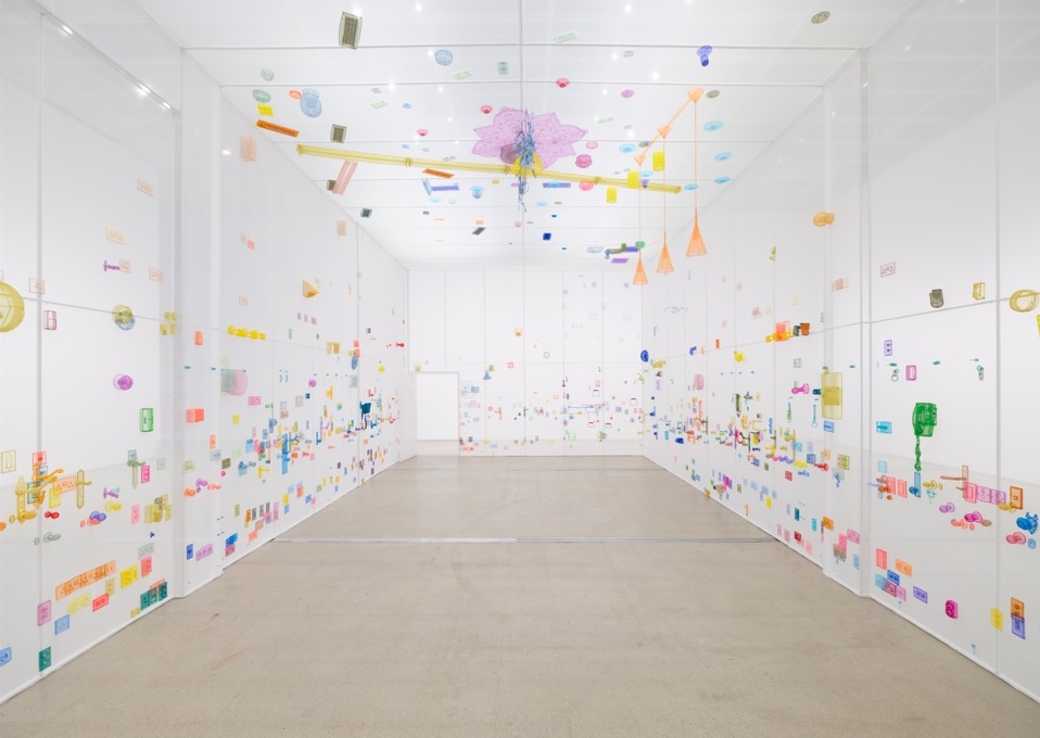 An installation view of a gallery space with white walls covered in hundreds of small, brightly colored objects and a hanging mobile sculpture suspended from the ceiling.