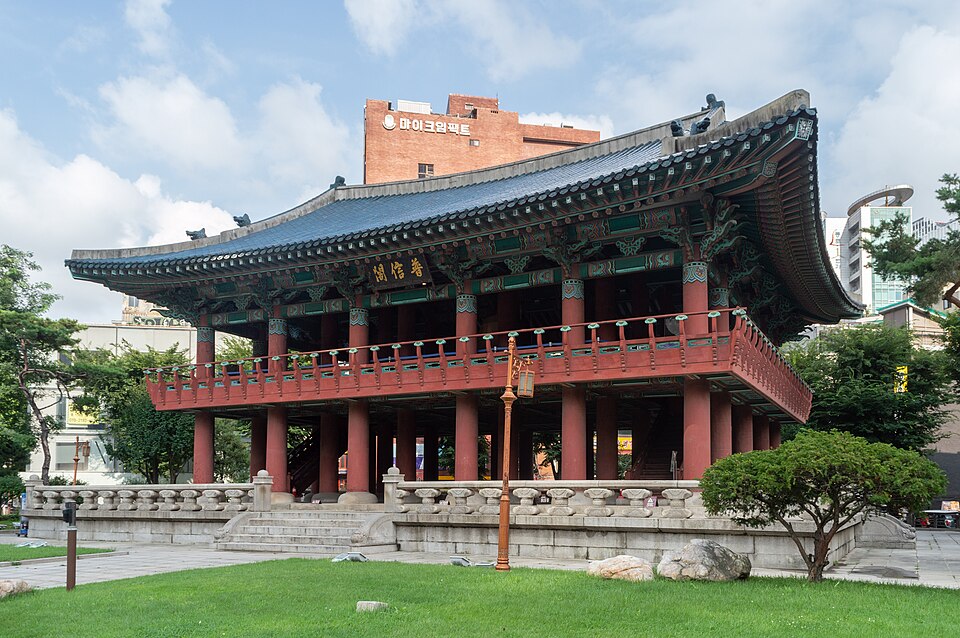 A traditional red and green Korean pavilion called Bosingak, with a large bronze bell visible beneath its roof, stands against a clear blue sky with modern city buildings in the background.