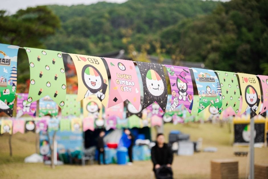 Colorful pennant banners featuring a cartoon gimbap mascot line an outdoor festival area with tents and trees in the background.