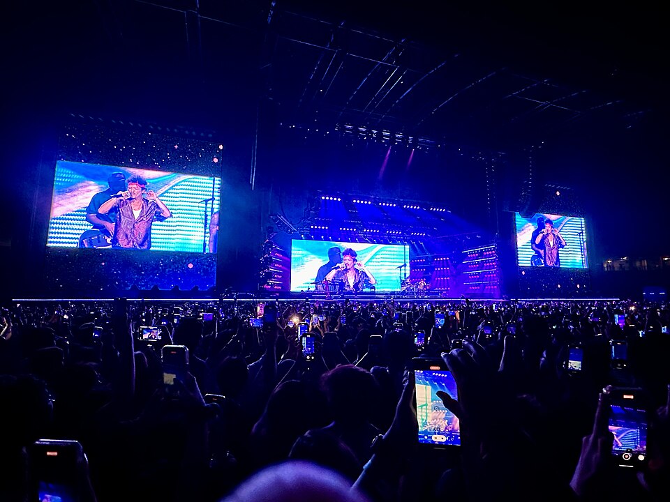 A stadium concert screen shows a close-up of a singer holding a microphone, flanked by band members performing on a runway stage, with the crowd's raised phones visible in the foreground.