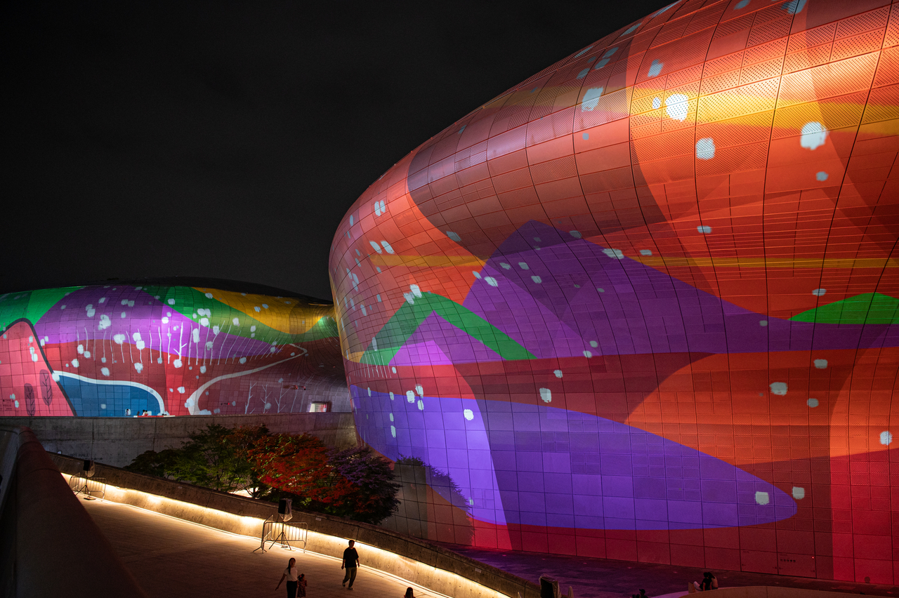Colorful abstract artwork resembling mountain shapes is projected at night onto the curved, tiled facade of Dongdaemun Design Plaza as pedestrians walk along an illuminated path below.