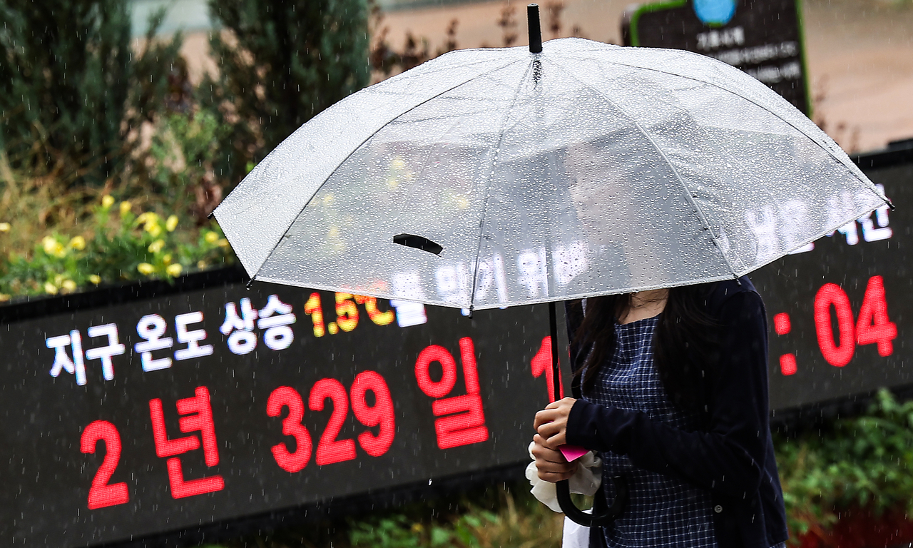 A person holds a clear umbrella in the rain while walking past an illuminated climate clock display.