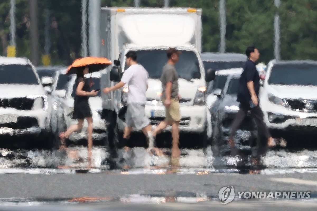 Blurred by heat haze, several pedestrians cross a road lined with parked vehicles, one shielding herself with an orange umbrella under bright sunlight.