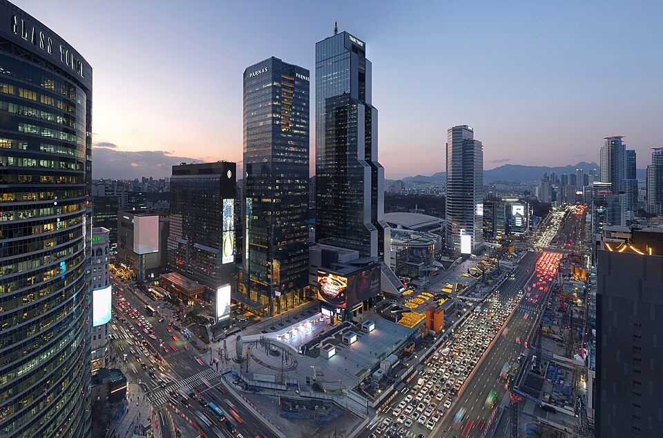 Aerial night view of a busy multi-lane intersection in Gangnam, Seoul, lined with illuminated skyscrapers and dense traffic.
