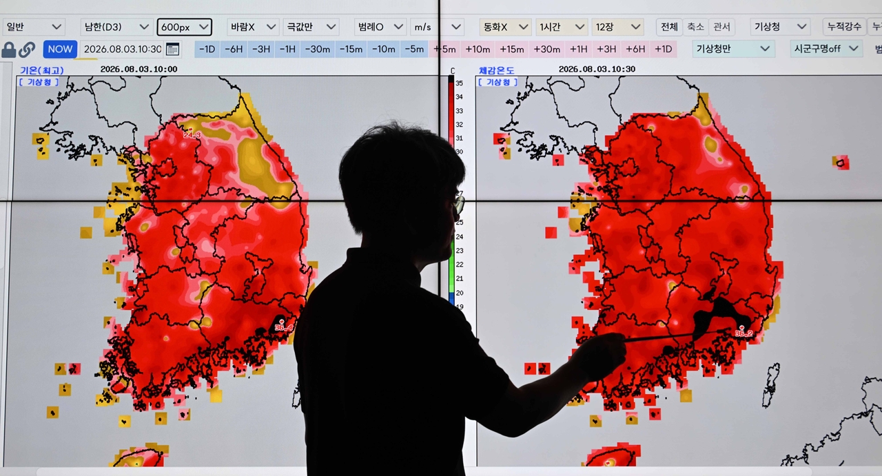A silhouetted man points at large screens displaying red-highlighted heat and temperature maps of the Korean peninsula inside a weather agency office.