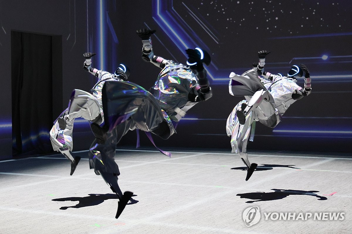 Three humanoid robots in shiny silver costumes with glowing wrist and head accents perform acrobatic flips on a stage lit with blue circuit-line patterns and a starry backdrop.