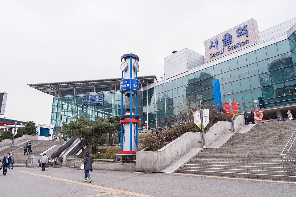 The exterior of Seoul Station is shown with its glass facade, an illuminated sign reading "서울역 Seoul Station," and a tall clock tower with red and blue decorative rings in front of the entrance stairs.