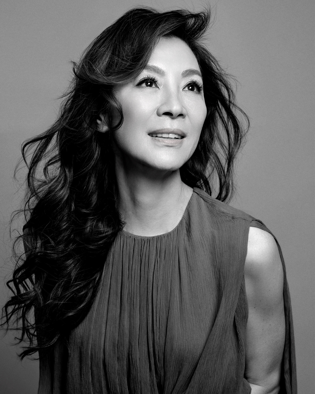 Black-and-white portrait of a woman with long wavy dark hair wearing a draped sleeveless top, looking upward and smiling slightly.