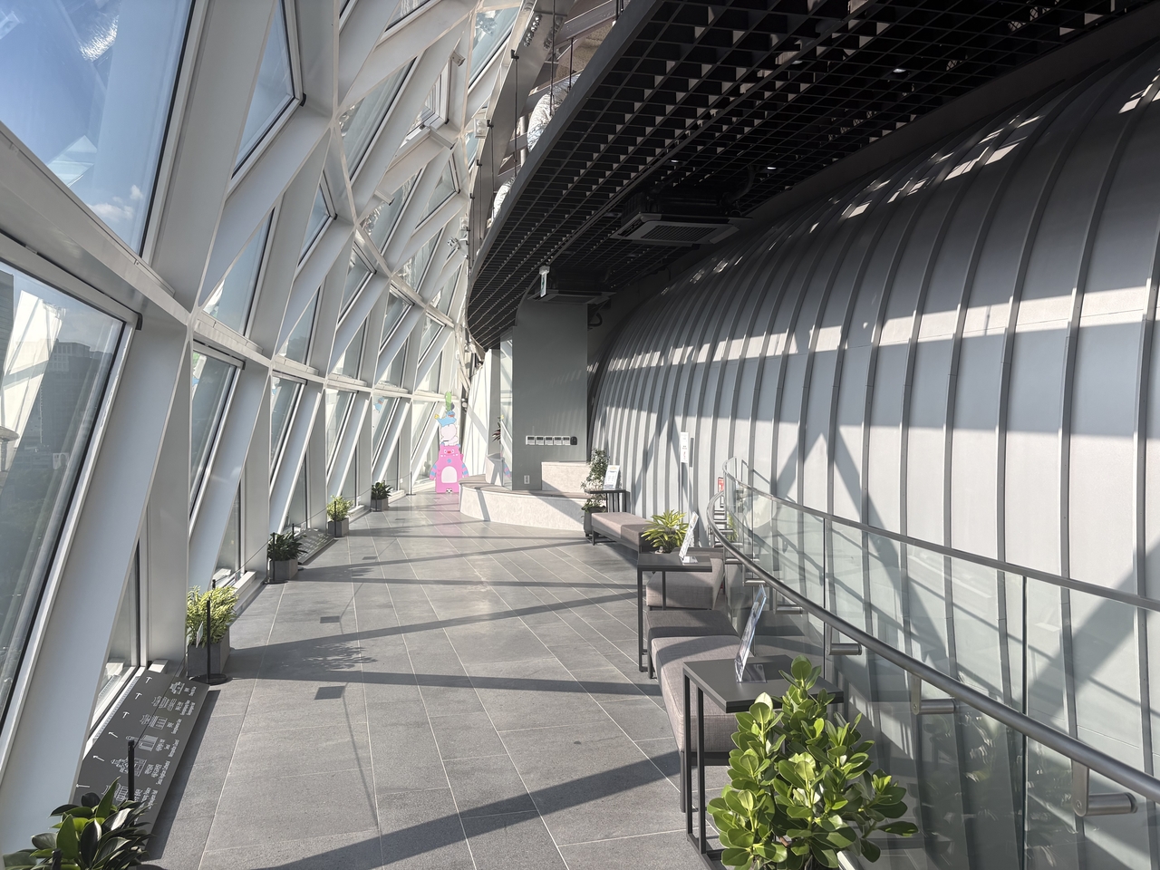 A curved glass-walled corridor with tiled flooring, potted plants and lounge seating inside an observation deck.