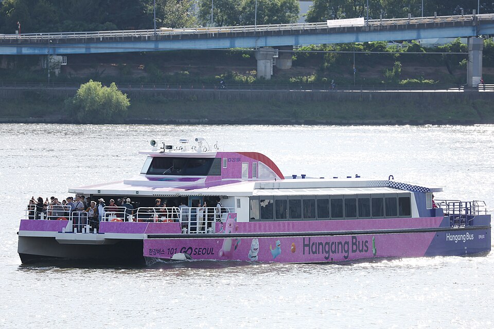 A pink and purple Hangang Bus river ferry, marked '101 GO SEOUL' and 'Hangang Bus', carries passengers on its open deck as it cruises the Han River beneath a blue overpass bridge.