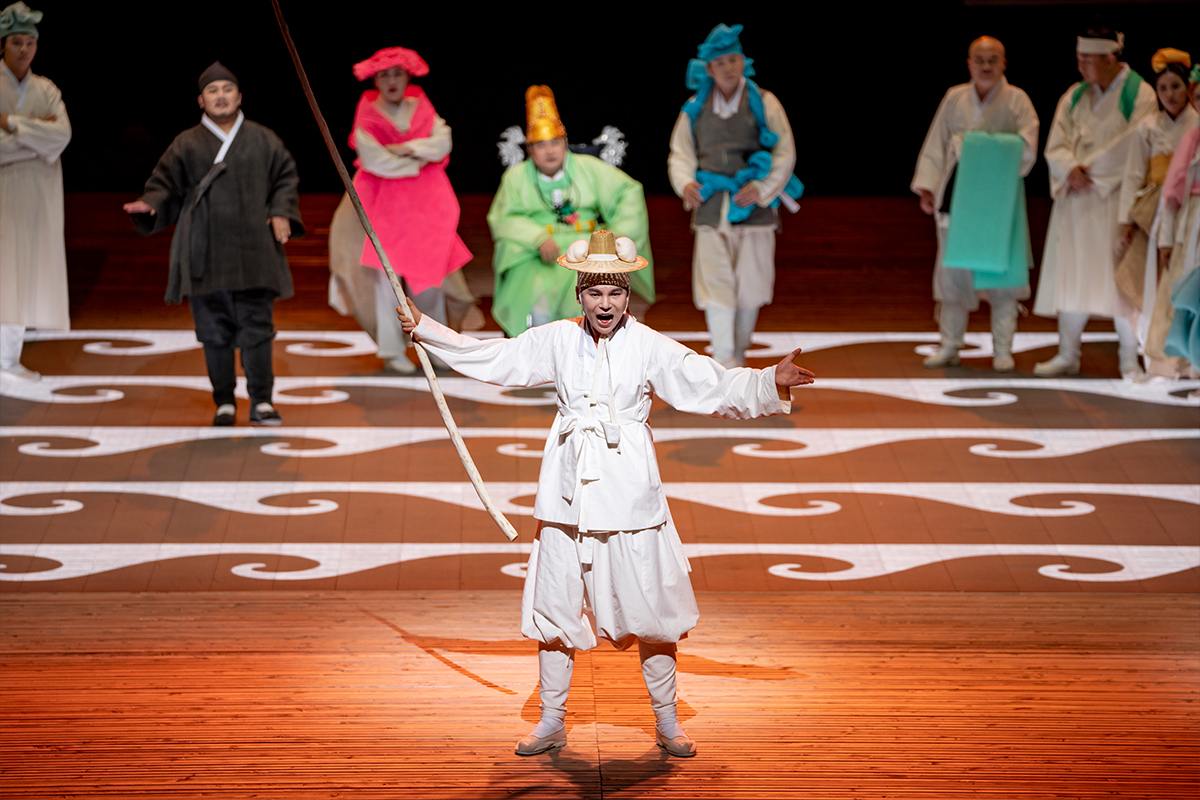 A performer dressed in white traditional Korean clothing raises a long staff and gestures on a wooden stage, with a row of colorfully costumed cast members standing behind him.
