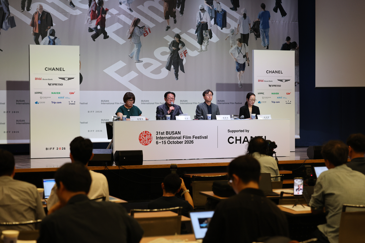 A man speaks into a microphone at a press conference table with three other panelists seated behind name placards, in front of a large backdrop reading Busan International Film Festival, as photographers and reporters watch from the audience.