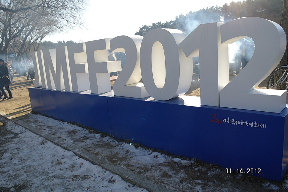 Large white block letters spelling out a film festival name and year stand on a blue base outdoors near bare trees, with people walking nearby and smoke rising in the background.