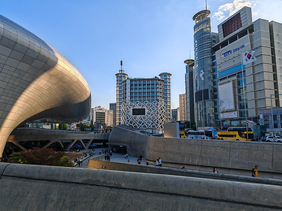Daytime view of the curved metallic exterior of Dongdaemun Design Plaza with surrounding city buildings and tour buses parked nearby.