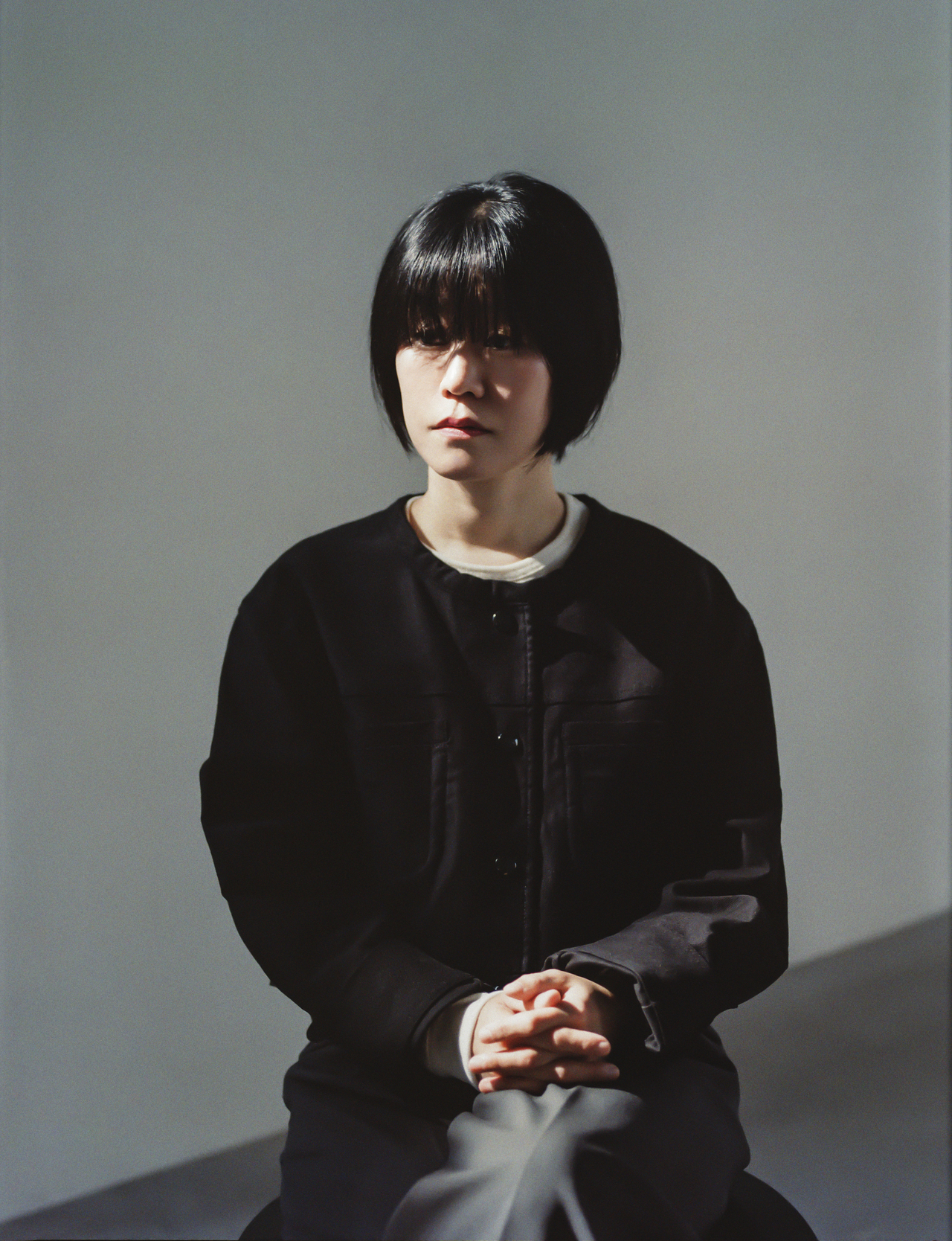 A woman with a short black bob haircut sits with hands clasped, wearing a black jacket over a light top, against a plain gray background.