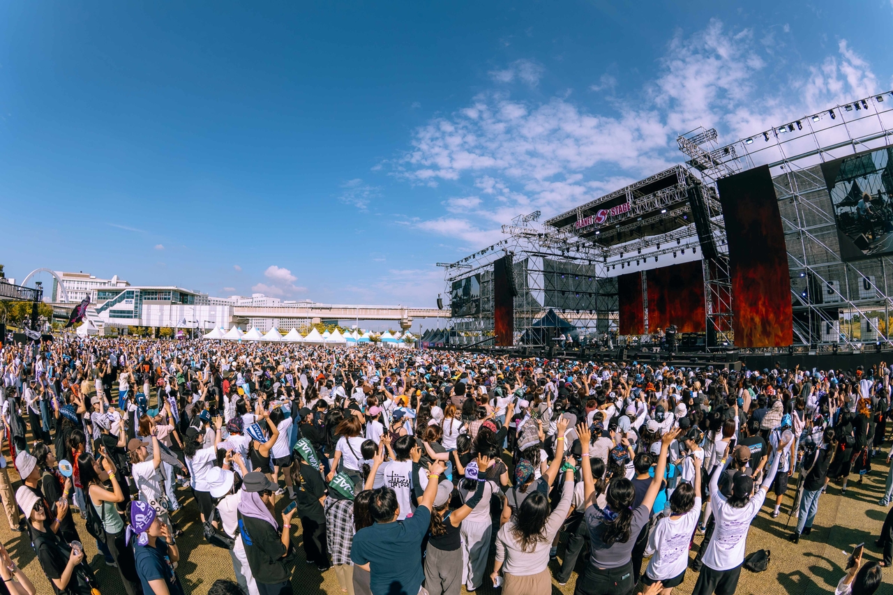 A large crowd of festival attendees with raised hands faces an outdoor concert stage under a blue sky with scattered clouds.