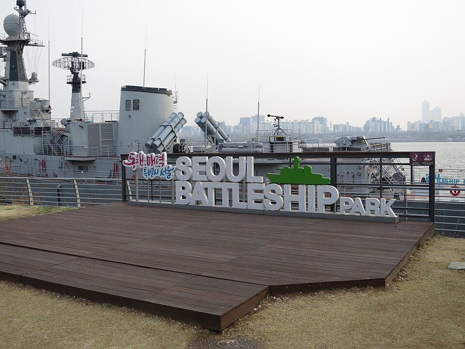 A sign reading "Seoul Battleship Park" stands on a wooden platform in front of a large gray naval warship docked at the riverside park, with the Seoul skyline visible in the hazy background.