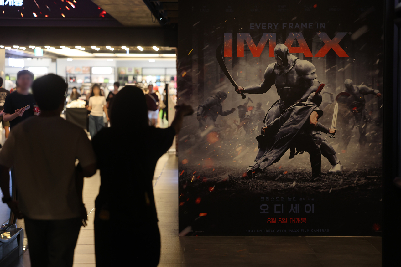 A movie theater poster for Christopher Nolan's film 'The Odyssey' displayed in an IMAX cinema lobby, with silhouettes of two people walking past and other moviegoers visible in the background.