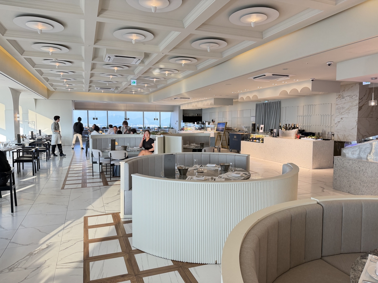A modern restaurant dining area with round upholstered booths, marble flooring, and floor-to-ceiling windows overlooking a city skyline.