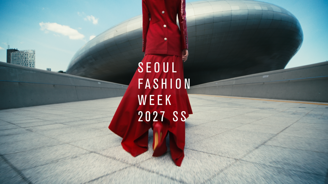 A person dressed in a red tailored jacket and long red skirt with red heels stands on a paved plaza in front of the large curved metallic exterior of the Dongdaemun Design Plaza under a blue sky.