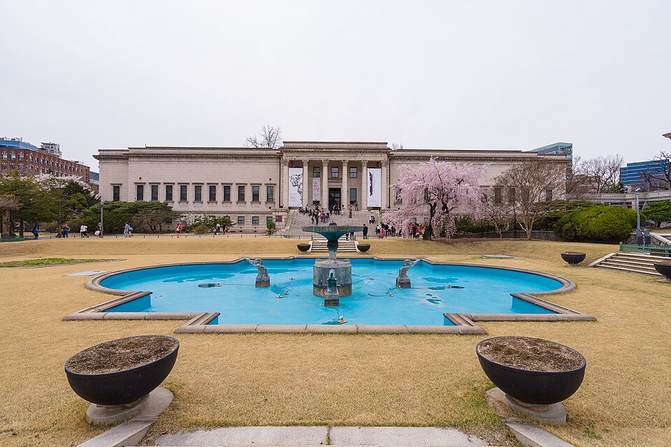 A grand neoclassical stone building with columned portico stands behind a green lawn and a circular fountain under a partly cloudy sky.