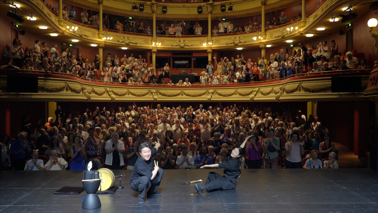 In an ornate multi-tiered theater packed with applauding spectators, a man crouches on stage making a peace sign while a woman sits nearby with her arm raised, a drum and stand beside them.
