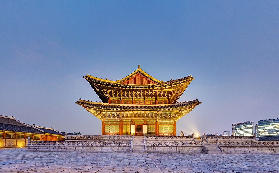A two-tiered traditional Korean palace hall, illuminated at night with warm lighting on its ornate wooden columns and eaves, seen from an angled side view against a dark sky.