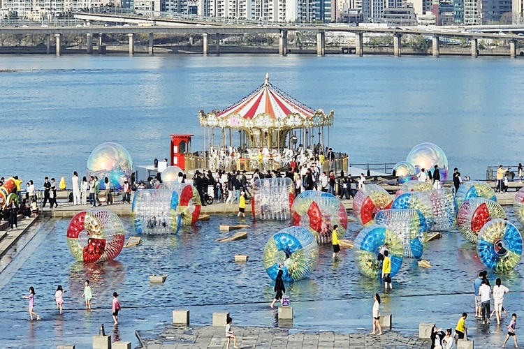 Crowds walk and play among large inflatable water rolling balls on the Han River waterfront in Seoul, with a colorful carousel and city skyline in the background.