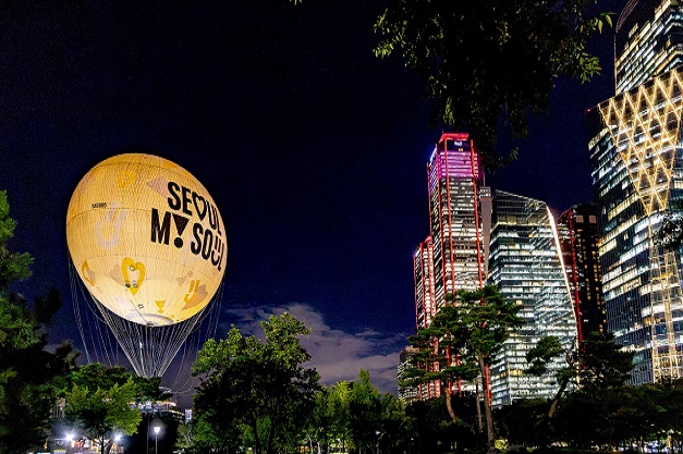 A glowing golden hot-air balloon reading "Seoul My Soul" floats above trees at night beside illuminated high-rise skyscrapers.