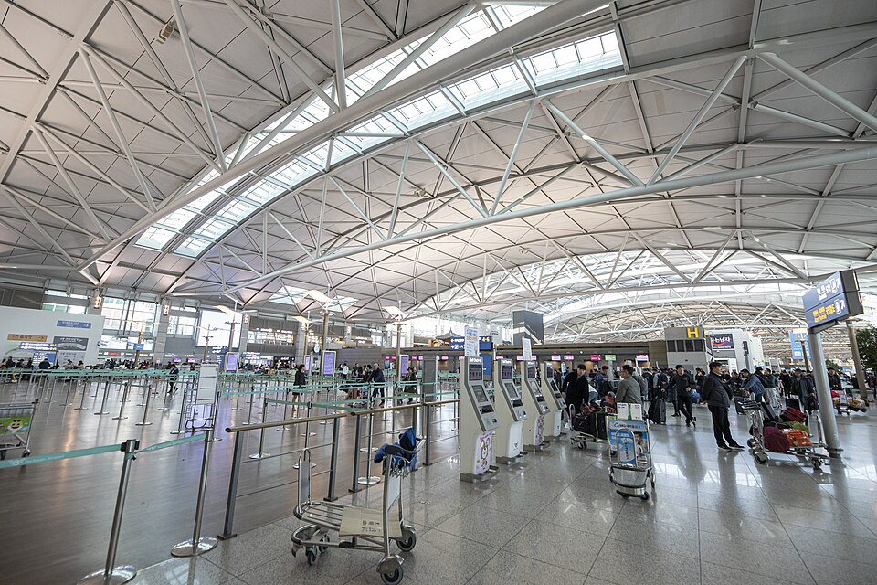 A wide, brightly lit view of Incheon International Airport Terminal 1's departure hall, showing rows of check-in counters, self-service kiosks, luggage carts and a few travelers beneath a curved metal-and-glass roof.