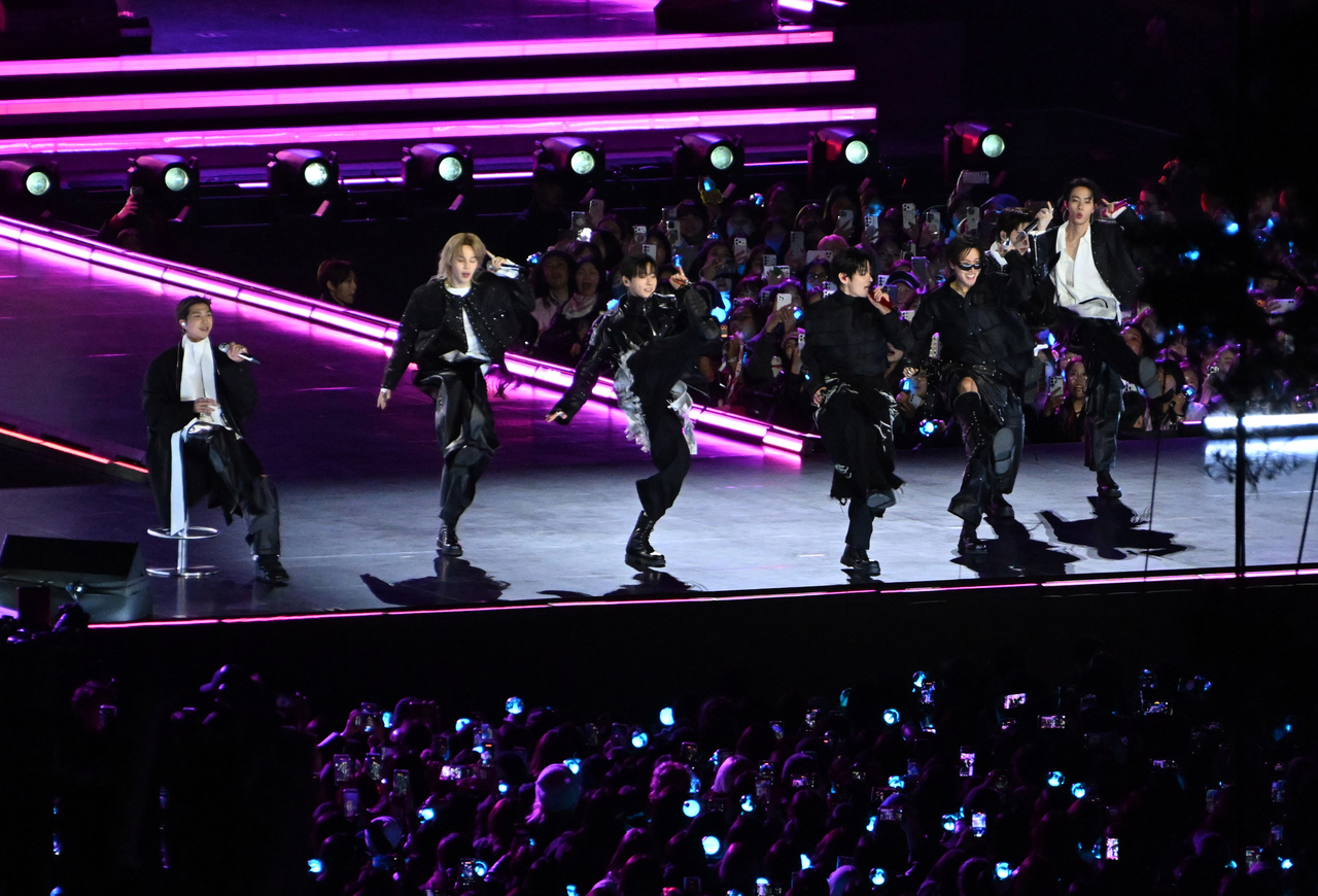 Six performers in black stage outfits dance and sing on a concert stage lit with pink and purple lights, with a large crowd holding up phones in the background.