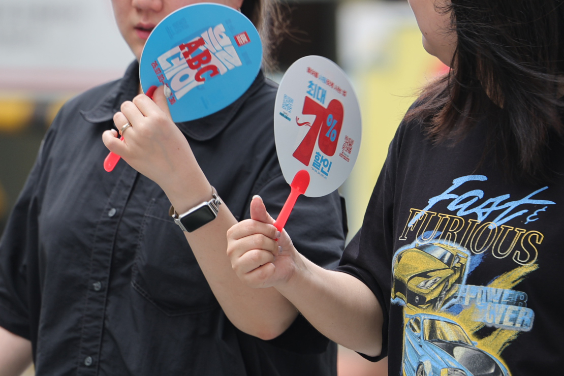 Two people hold red-handled promotional fans, one printed with blue lettering and another advertising a percentage discount, partially covering their faces as they stand close together outdoors.