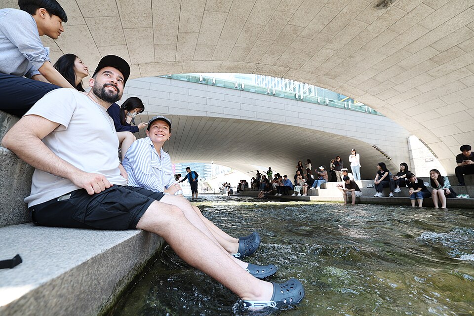 People sit along a stone ledge with their feet dangling in the shallow water of Cheonggyecheon stream under a concrete bridge, while others sit and stand along the opposite bank.