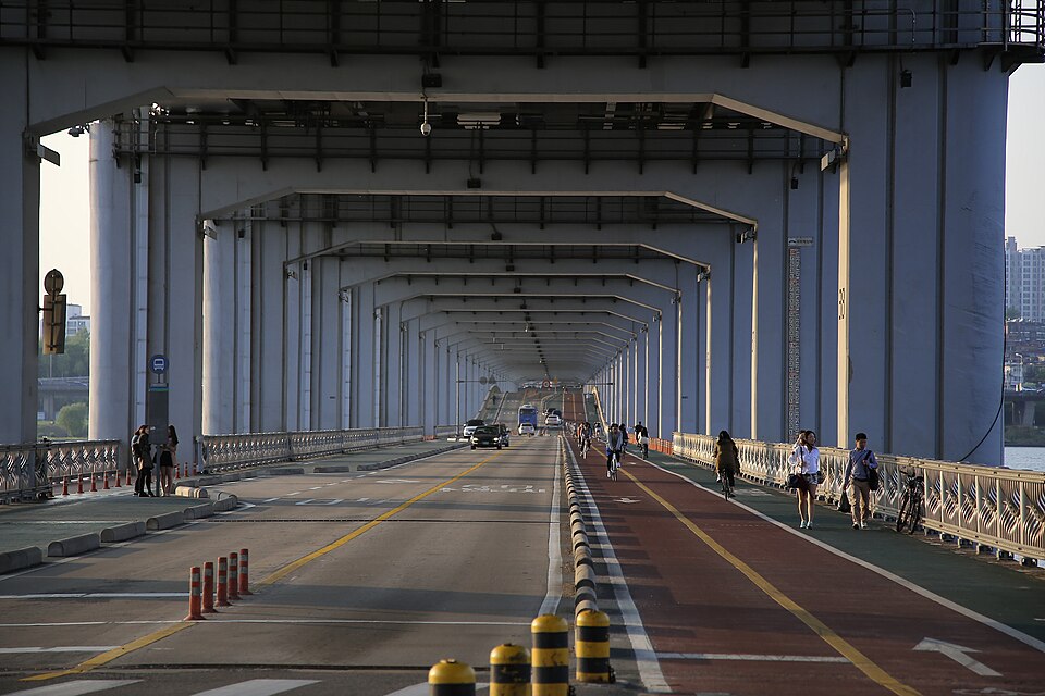 Pedestrians and cyclists use marked lanes on Jamsugyo Bridge in Seoul, with a few cars in the roadway and the elevated Banpo Bridge structure overhead in late afternoon light.